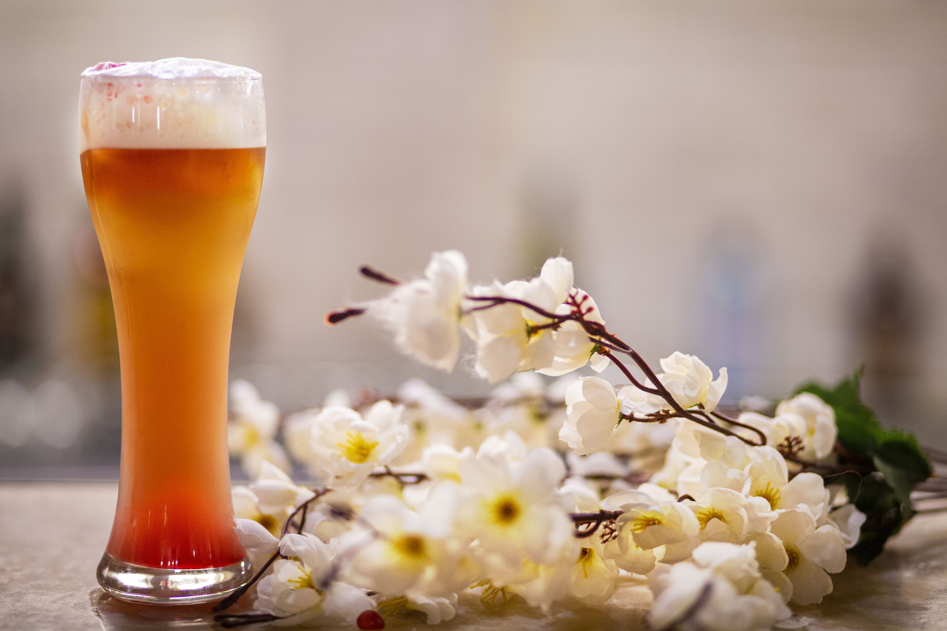 Un grand verre de bière, surmonté d’une mousse abondante, entouré de fleurs blanches et de feuilles vertes, le tout posé sur une surface réfléchissante.