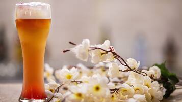 A tall glass of beer with a frothy head, surrounded by white flowers and green leaves on a reflective surface.
