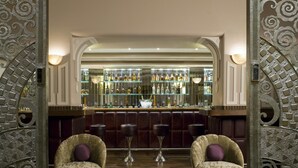 A bar area with a high bar counter, multiple shelves of bottles, and two cushioned armchairs with a decorative vase on the floor. - Ambassador, New Delhi - IHCL SeleQtions (New Delhi)