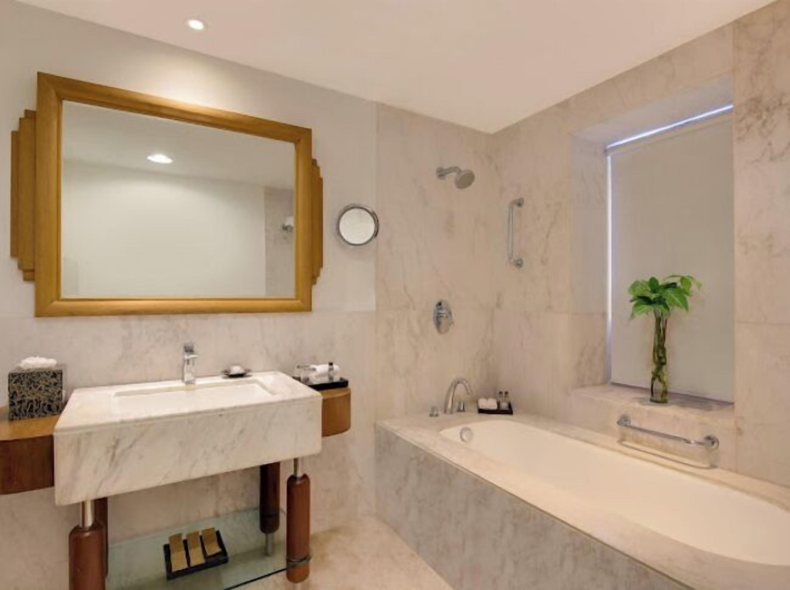 A modern bathroom with a large mirror, a sink, a bathtub, and a window with a plant.