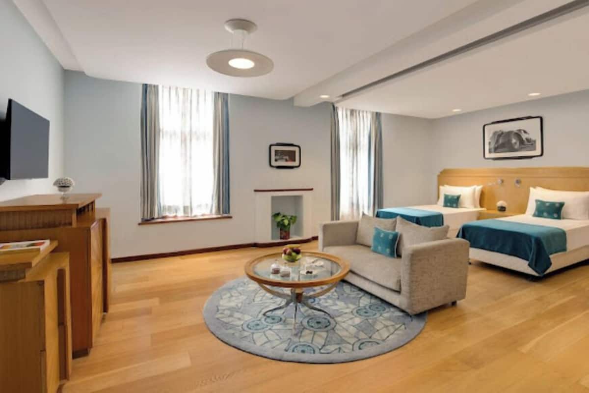 A modern hotel room with a sofa, a bed with blue bedding, a round coffee table, and a TV mounted on the wall.
