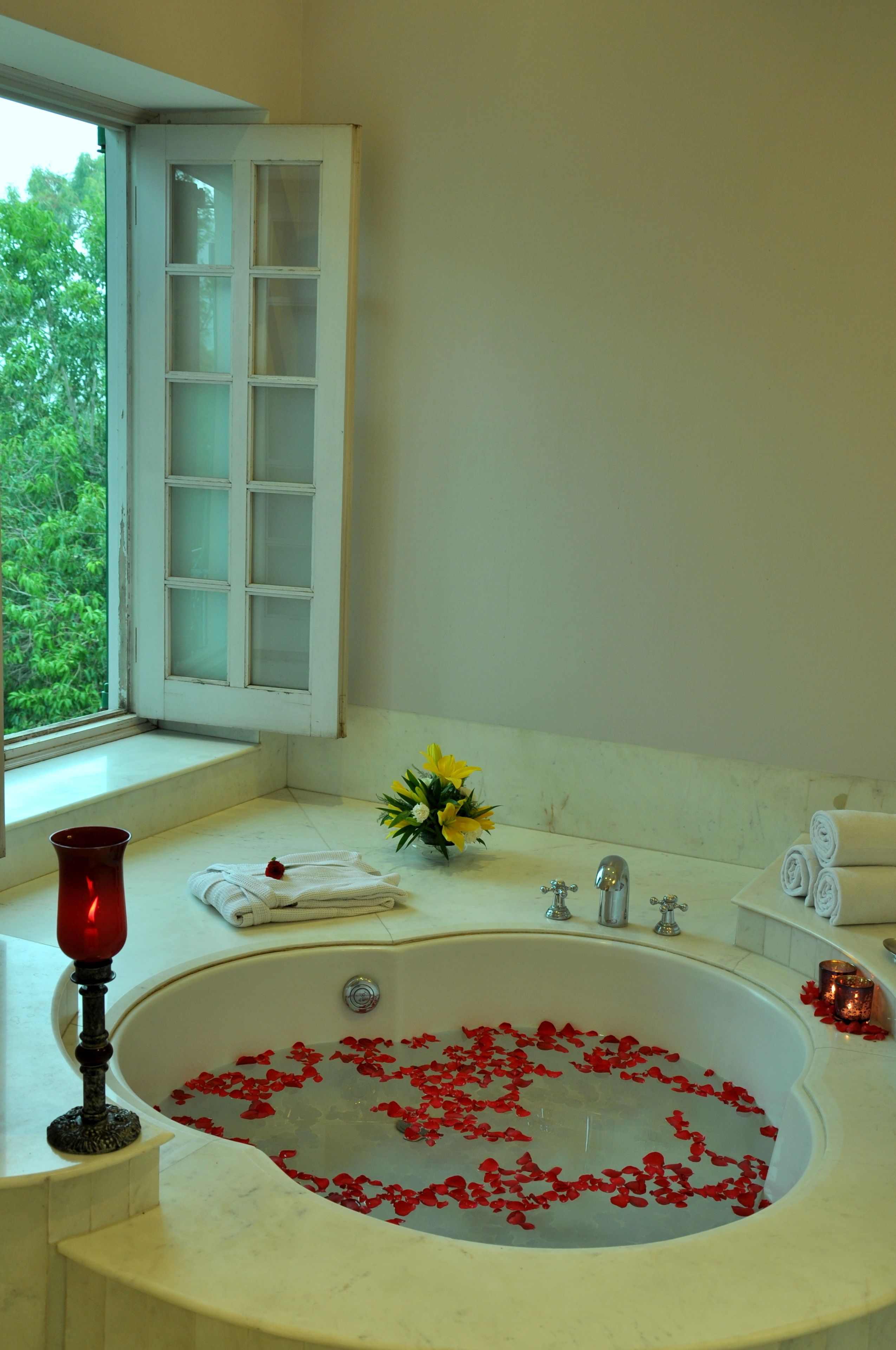 A bathroom with a bathtub filled with rose petals, a window with a view of greenery, and a vase with yellow flowers on the counter.