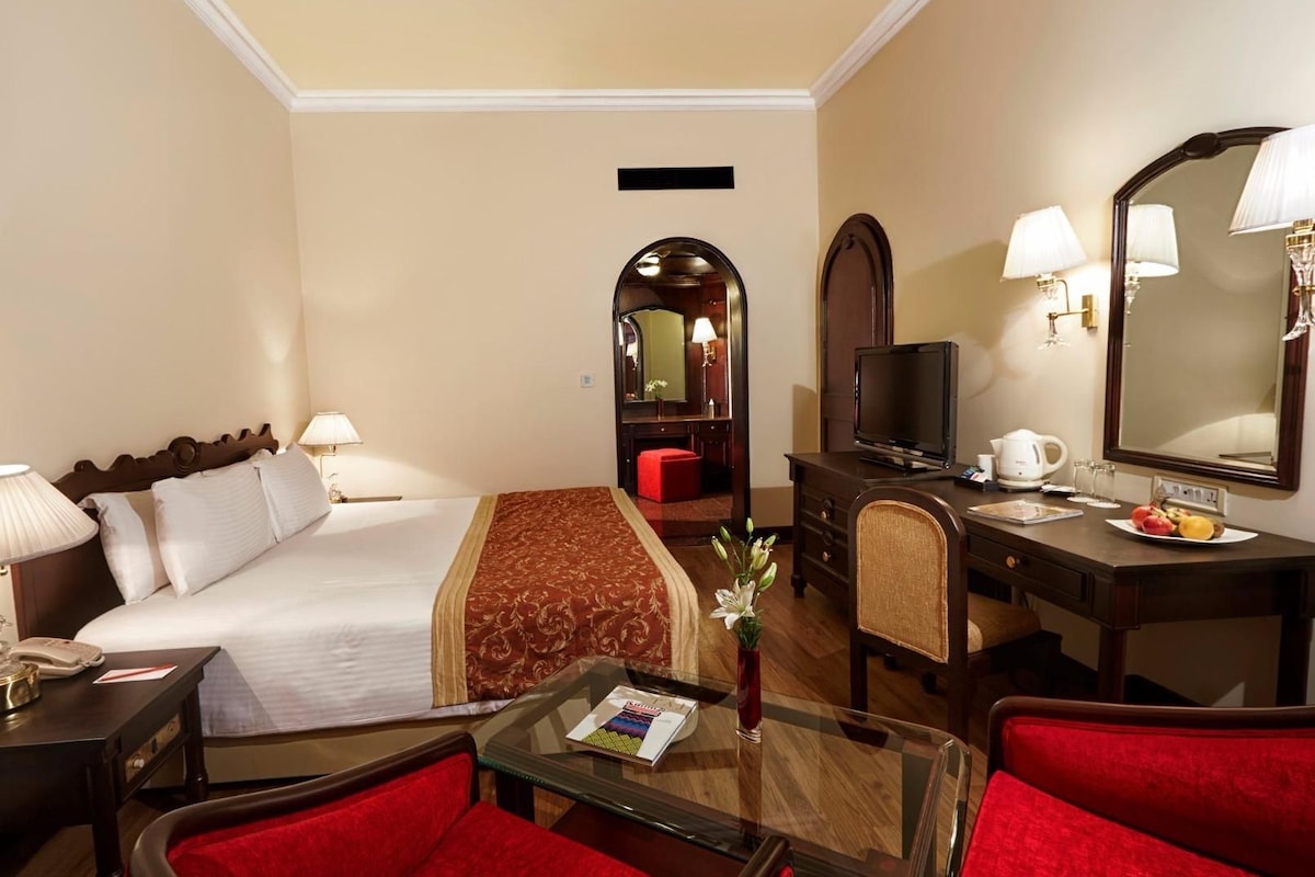 Una habitación de hotel con cama, escritorio, televisión, espejo y una silla roja.