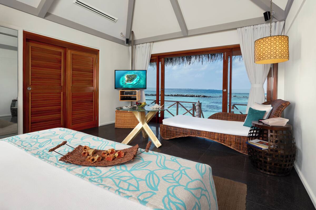 A bedroom with a bed, a television, a wicker chair, a table with a basket, and a view of the ocean.