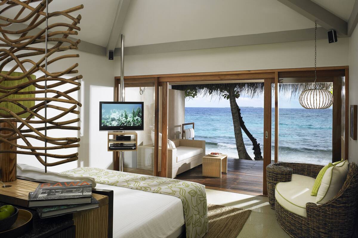 A bedroom with a bed, a TV, a wicker chair, and a view of the ocean.