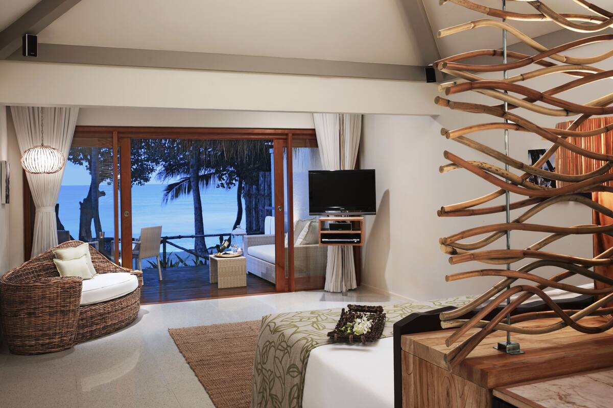 A modern hotel room with a balcony, wicker furniture, a flat-screen TV, and a unique wooden decor piece.