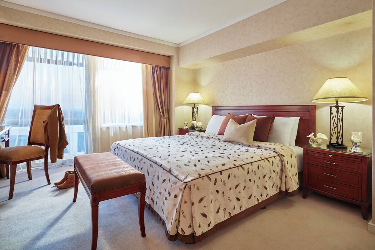 A hotel room with a large bed, a wooden bench, a chair, and a nightstand with lamps.