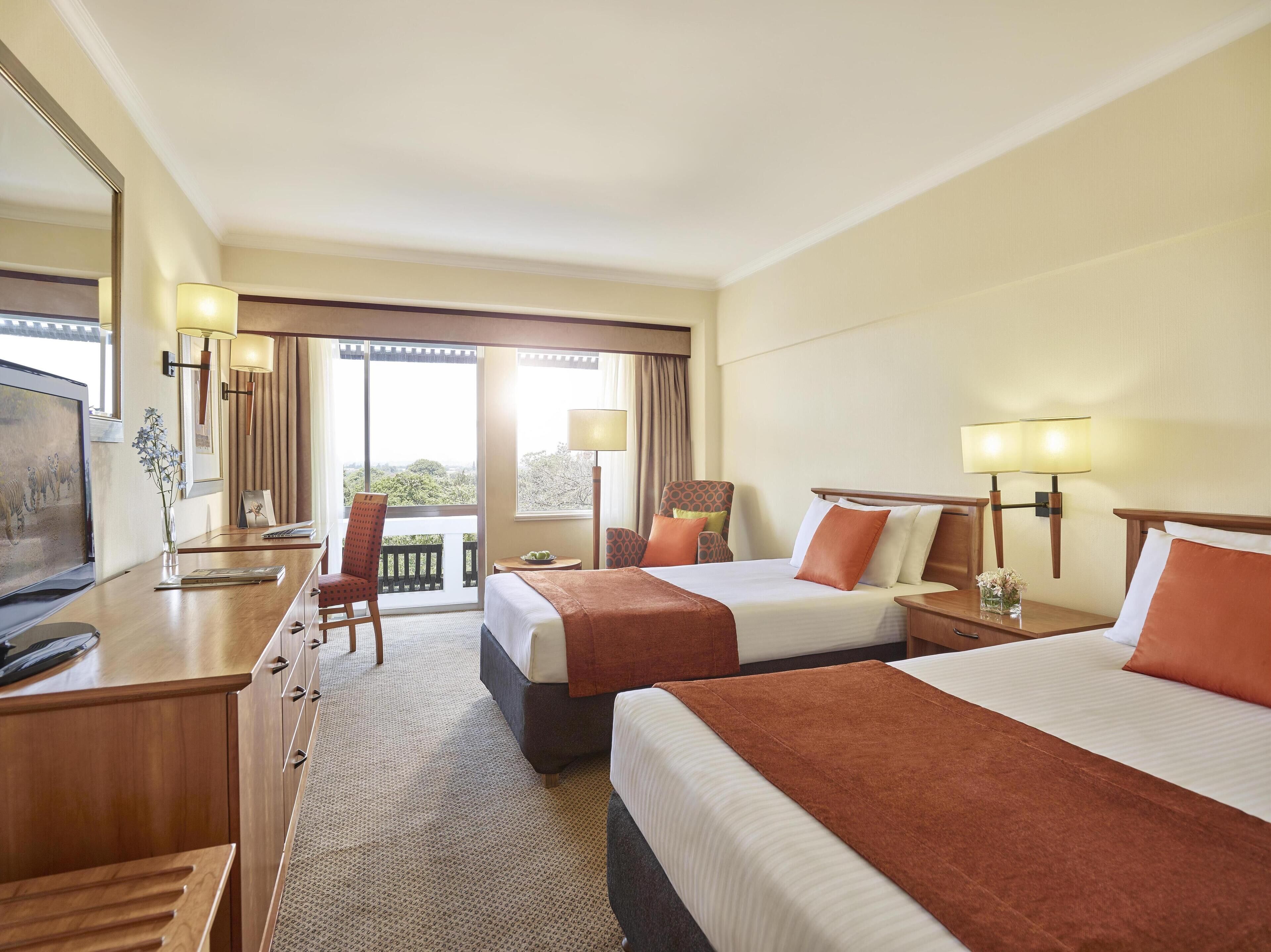 A hotel room with two beds, a desk, a TV, and a balcony with a view.