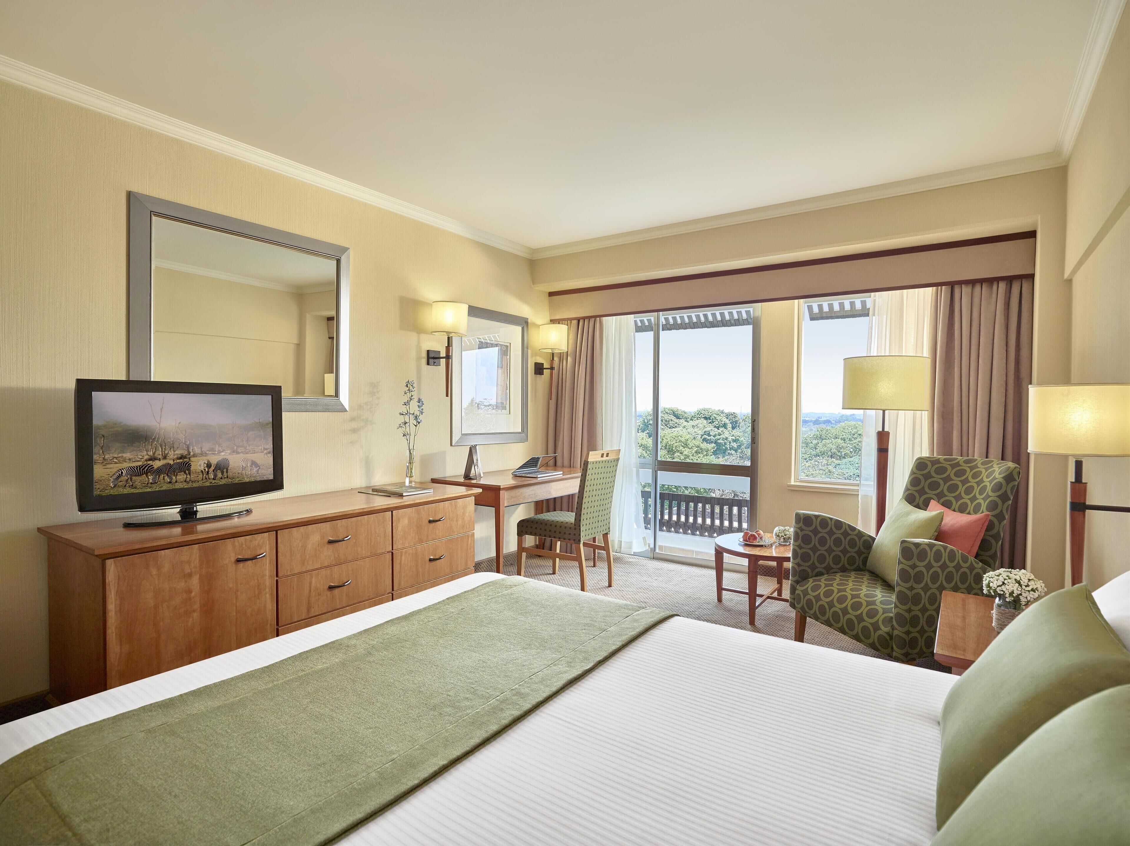 Habitación de hotel con cama, escritorio, silla, televisor y balcón con cortinas.