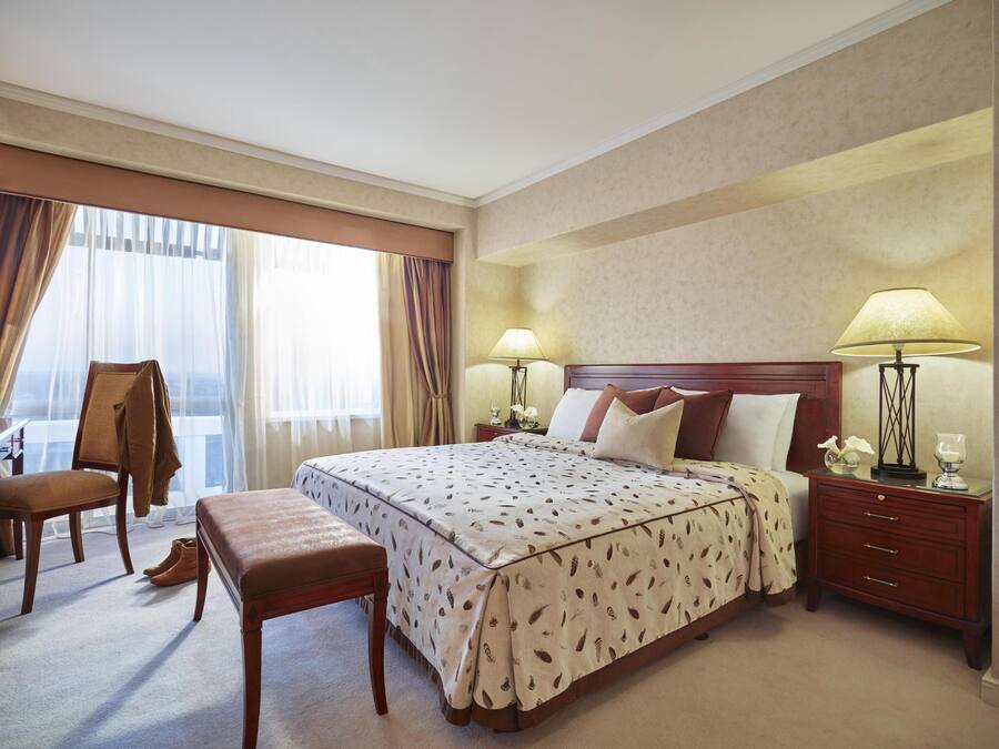 A hotel room with a large bed, two bedside tables with lamps, a chair, a bench, and a view of the city through large windows.