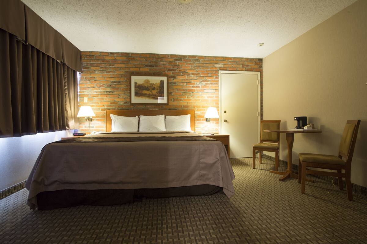Habitación de hotel con una cama grande, dos lámparas de noche, pared con diseño de ladrillo, una puerta, una mesita con cafetera y una silla.
