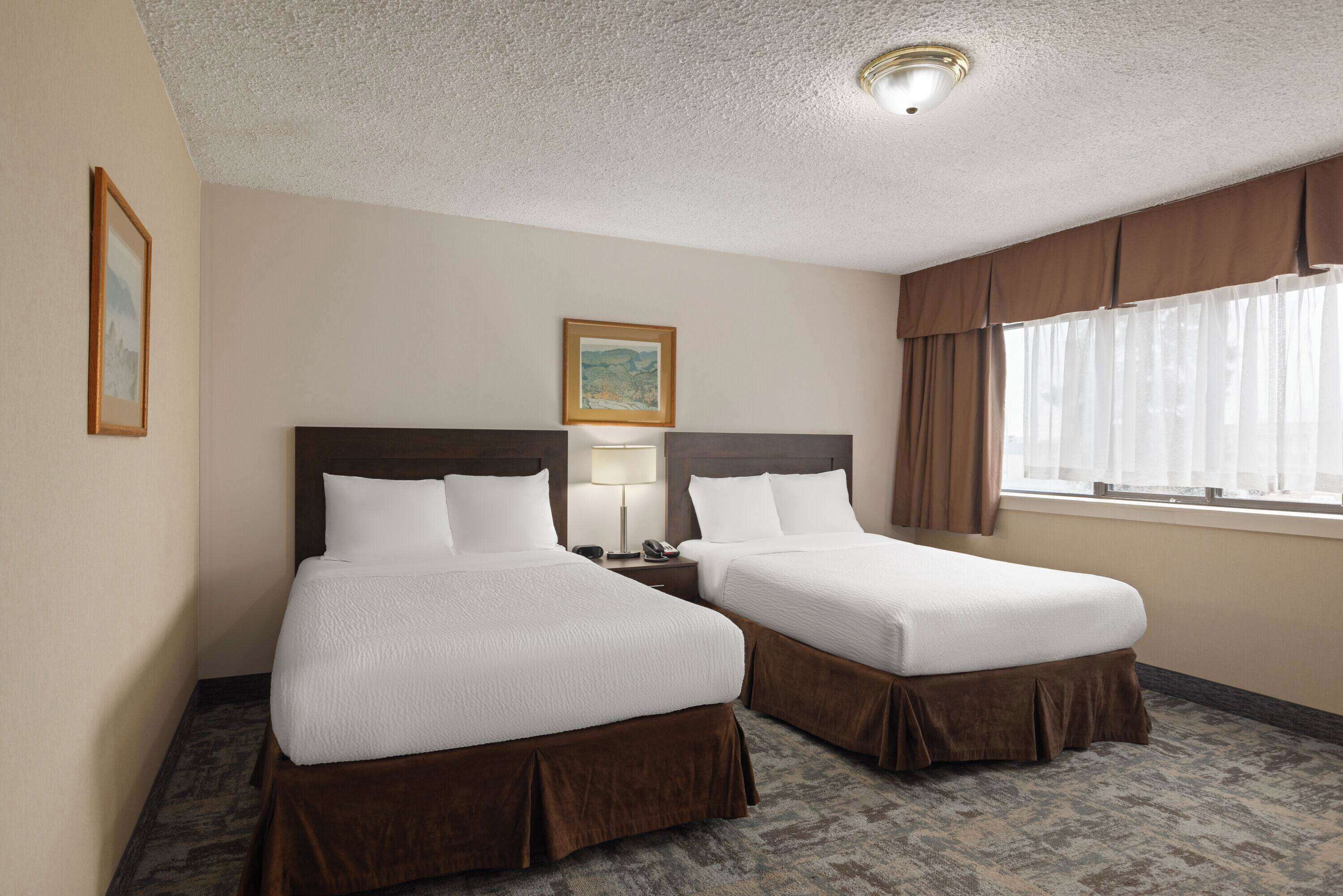 Quarto de hotel com duas camas de solteiro, mesa de cabeceira com abajur, quadro na parede e janela com cortinas.