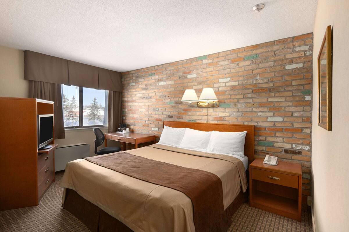 Habitación de hotel con pared de ladrillo, cama con cubrecama marrón, escritorio con silla, televisor y ventana con cortinas.