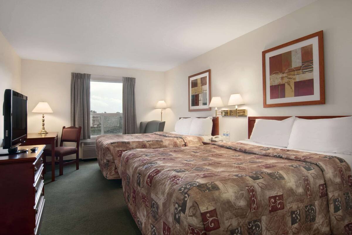 Habitación de hotel con dos camas, un escritorio con televisor y una silla. Hay dos lámparas en el escritorio y una ventana con cortinas.