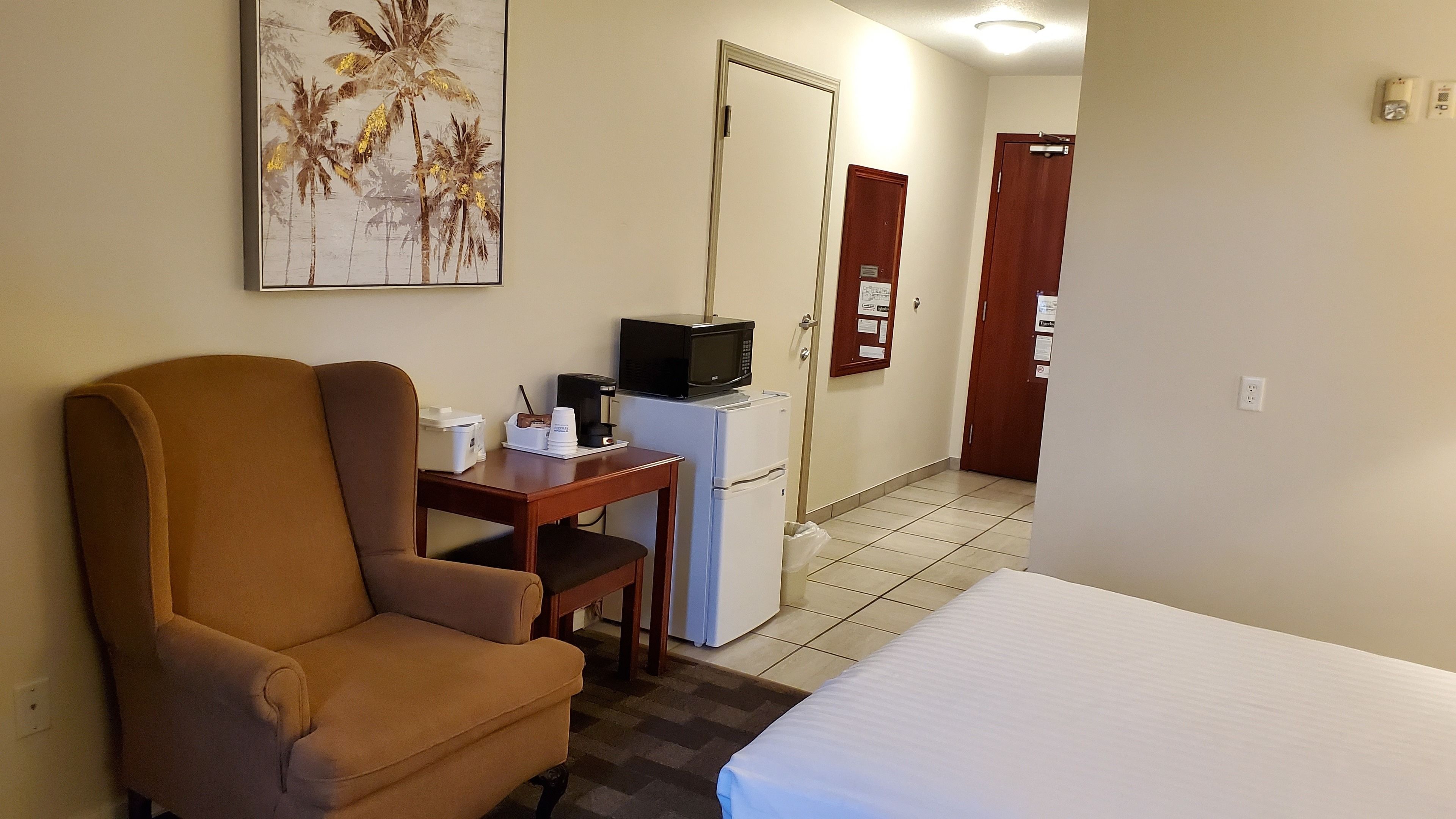 Quarto de hotel com uma cama, uma poltrona marrom, uma mesinha com micro-ondas e cafeteira, um frigorífico e uma porta com um painel de aviso.