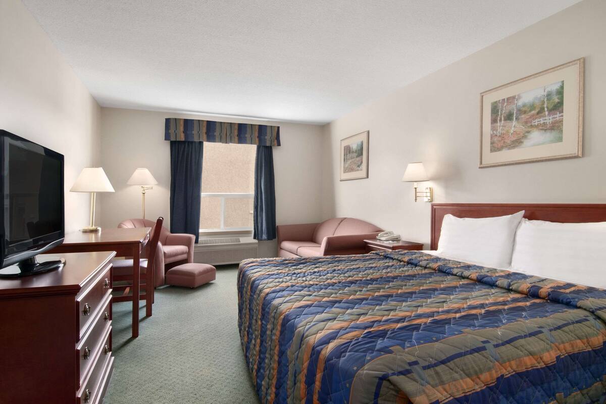 Habitación de hotel con cama, escritorio con televisor, silla, ventana con cortinas y un cuadro en la pared.