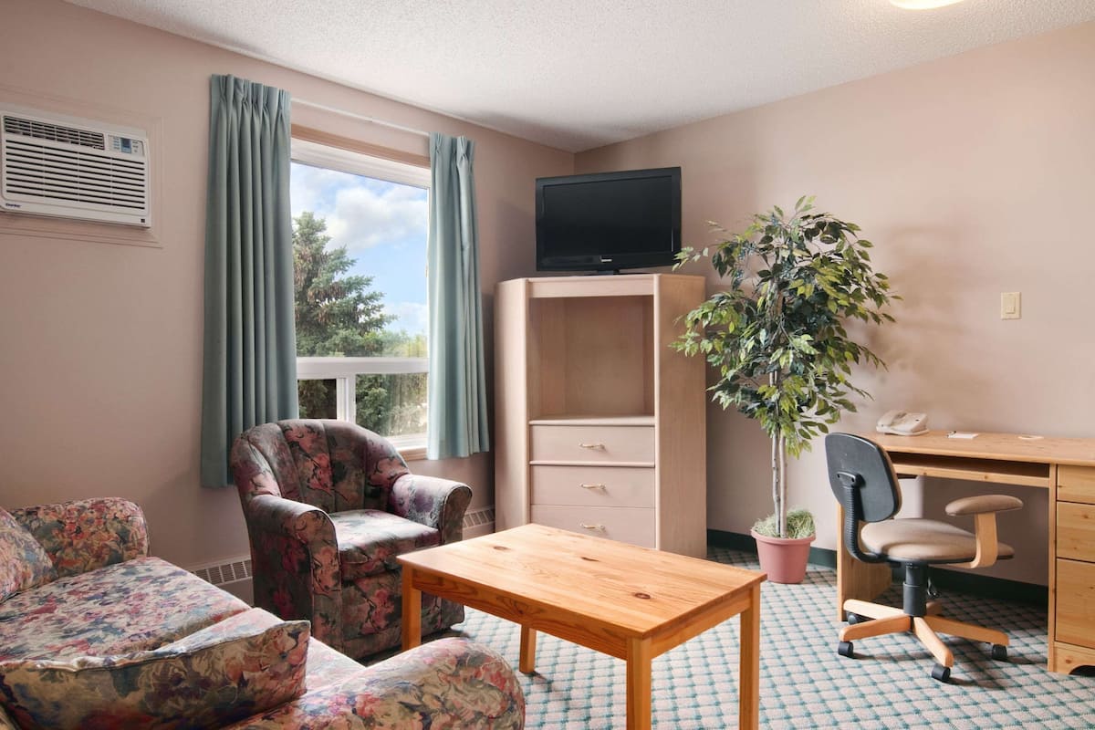 A room with a sofa, a wooden table, a desk with a chair, a television, a potted plant, and a window with curtains.