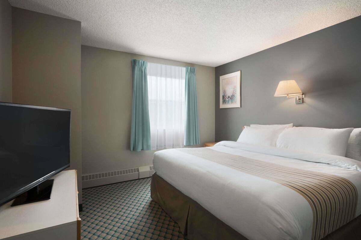 A hotel room with a bed, a television on a stand, a window with curtains, and a wall-mounted lamp.