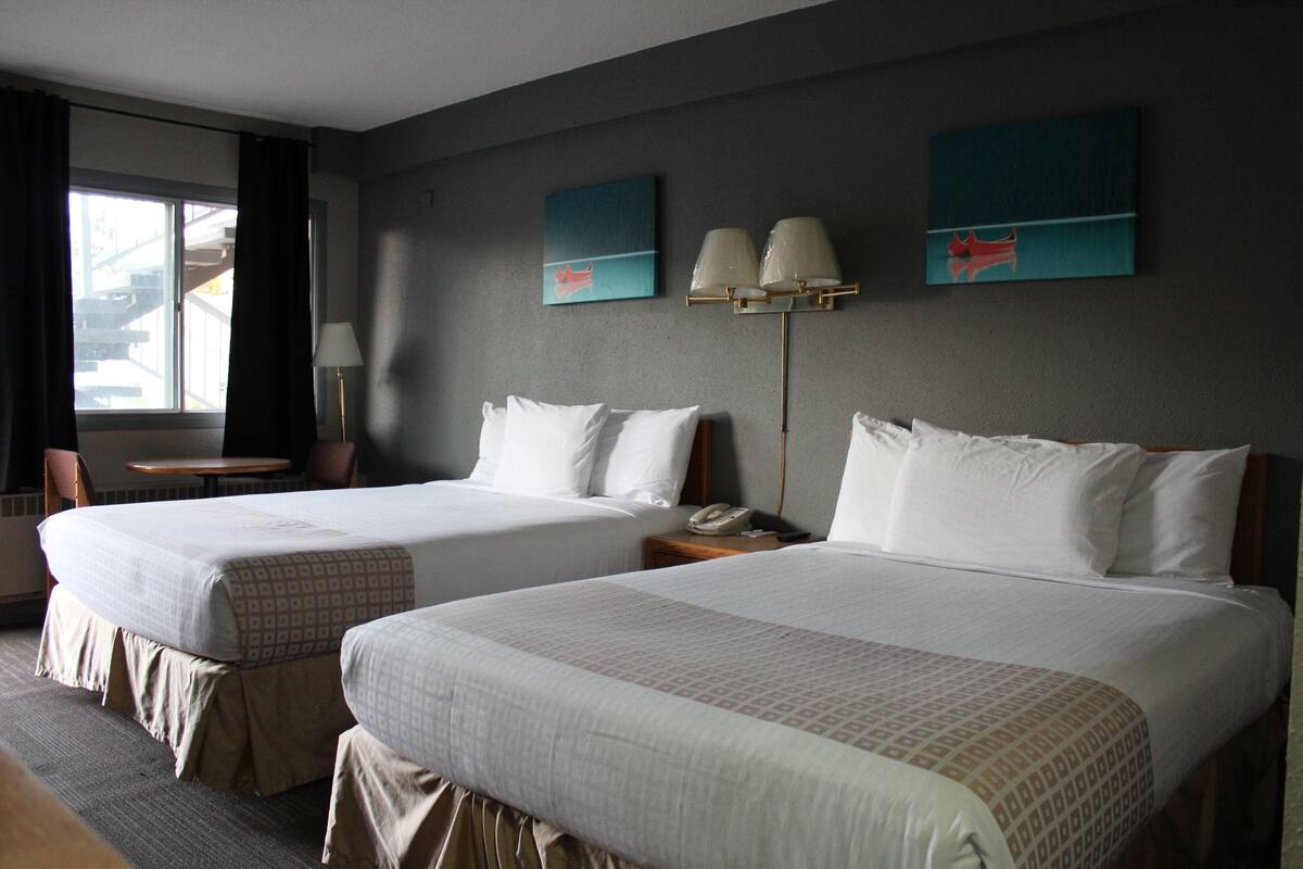 A hotel room with two beds, a desk, and a chair. There are two paintings on the wall and a lamp on the desk.