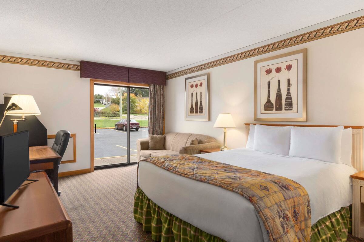 Hotelkamer met een bed, een bureau met een computer, een stoel, een lamp en uitzicht op de parkeerplaats.