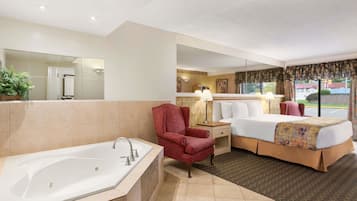 Habitación de hotel con una bañera grande, cama con colcha estampada, una silla y una mesita de noche con lámpara.