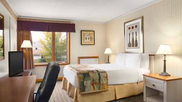 Ein Hotelzimmer mit einem Bett, zwei Nachttischen mit Lampen, einem Schreibtisch mit Computer, einem Sessel, einem Fenster mit Vorhängen und einem gerahmten Bild an der Wand.