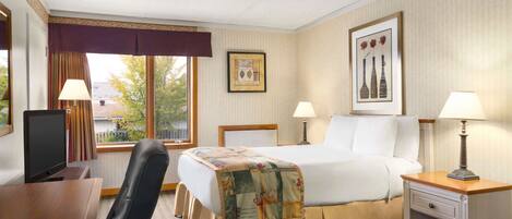 Habitación de hotel con cama, dos mesitas de noche con lámparas, un escritorio con ordenador, una silla, una ventana con cortinas y un cuadro en la pared.