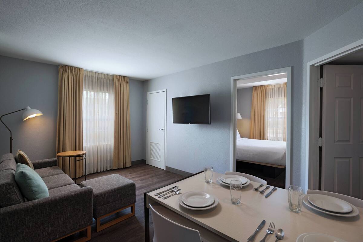 A hotel room with a dining area, a sofa, a TV mounted on the wall, and a door leading to another room.