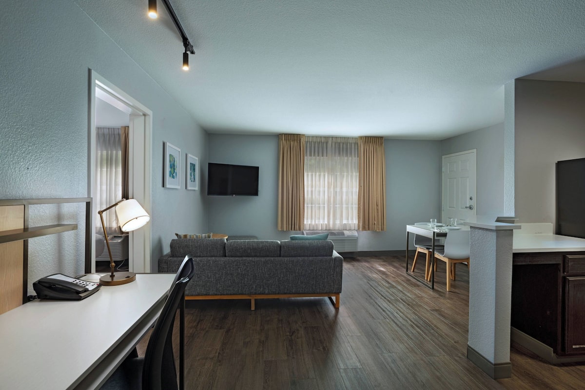 A modern hotel room with a sofa, a desk, a flat-screen TV, and a dining area.