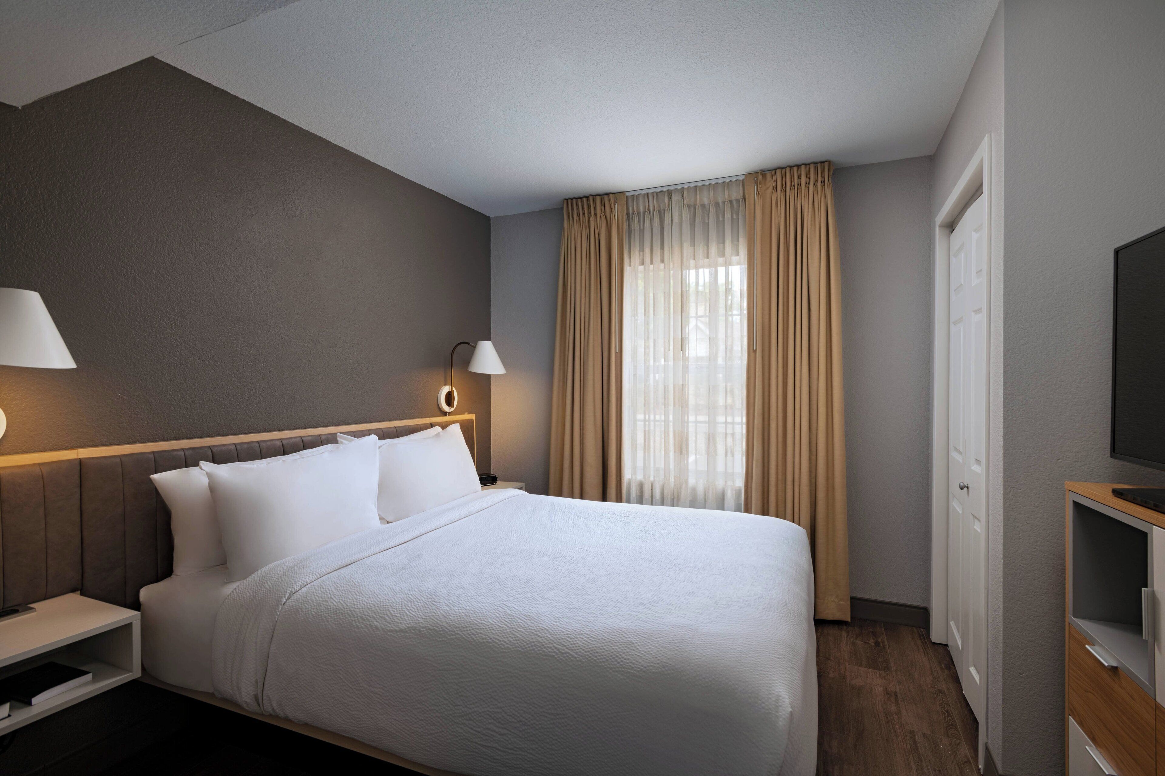 A hotel room with a large bed, two bedside lamps, a TV, and a window with curtains.