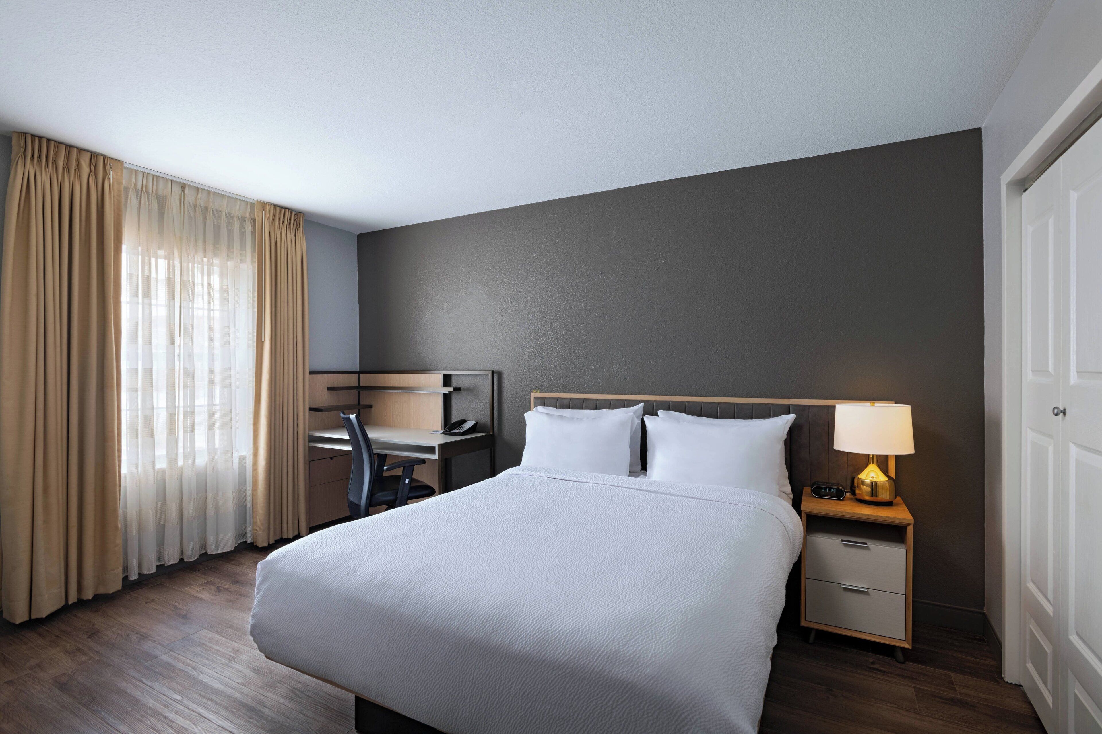 A hotel room with a bed, a desk, a chair, a nightstand with a lamp, and a door.