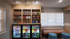 A seating area with a bench and two ottomans, a beverage refrigerator stocked with drinks, and a shelf with snacks and bottled water. - TownePlace Suites by Marriott Baton Rouge South (Baton Rouge)