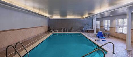 An indoor swimming pool with clear water, surrounded by tiled walls and a ceiling with recessed lighting. There are chairs and tables near the pool, and large windows on one side.