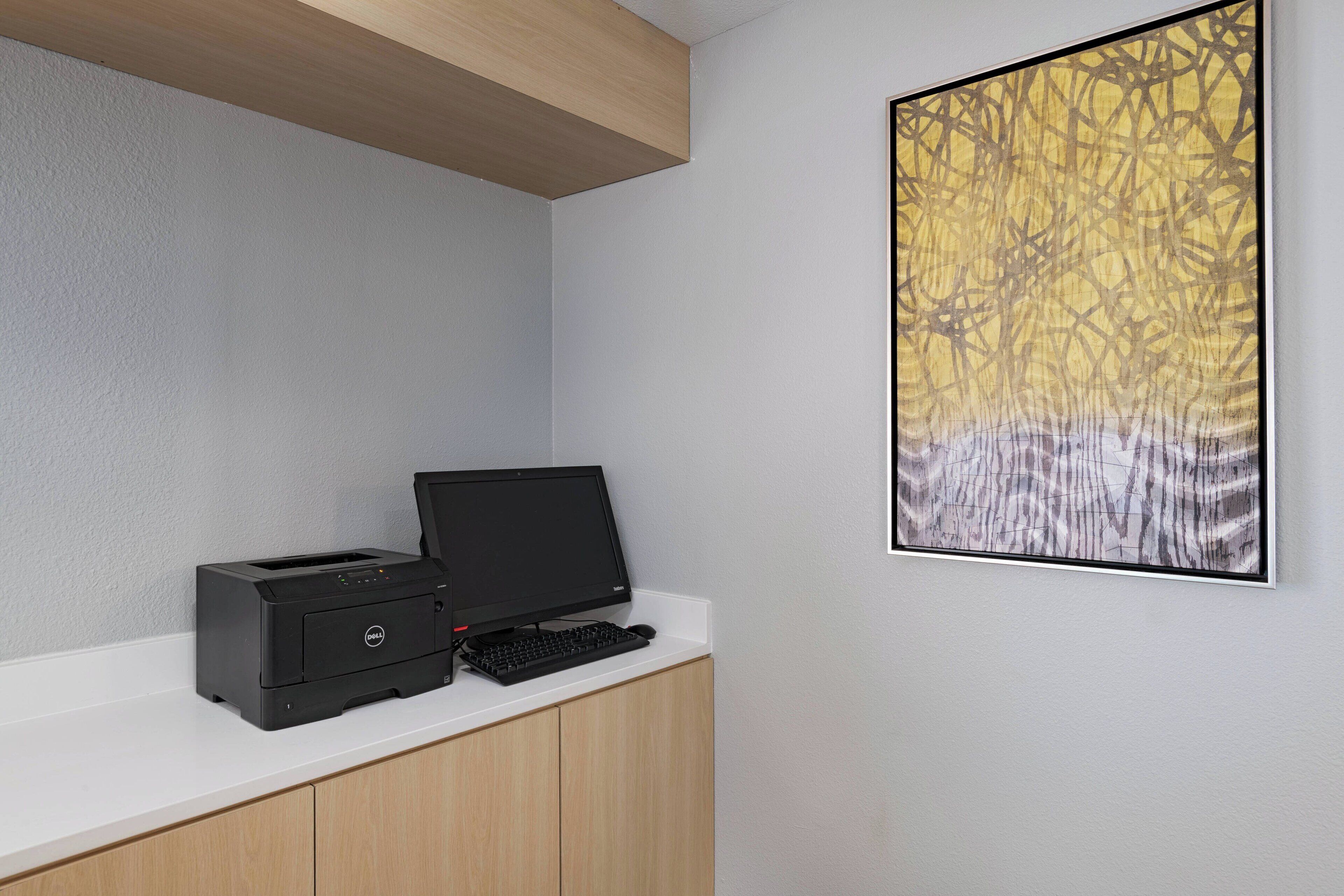 A workspace with a printer, keyboard, and monitor on a white desk against a light gray wall. A framed abstract painting is on the wall to the right.