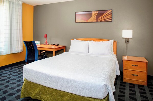 A hotel room with a bed, a desk, a chair, a nightstand, and a lamp.