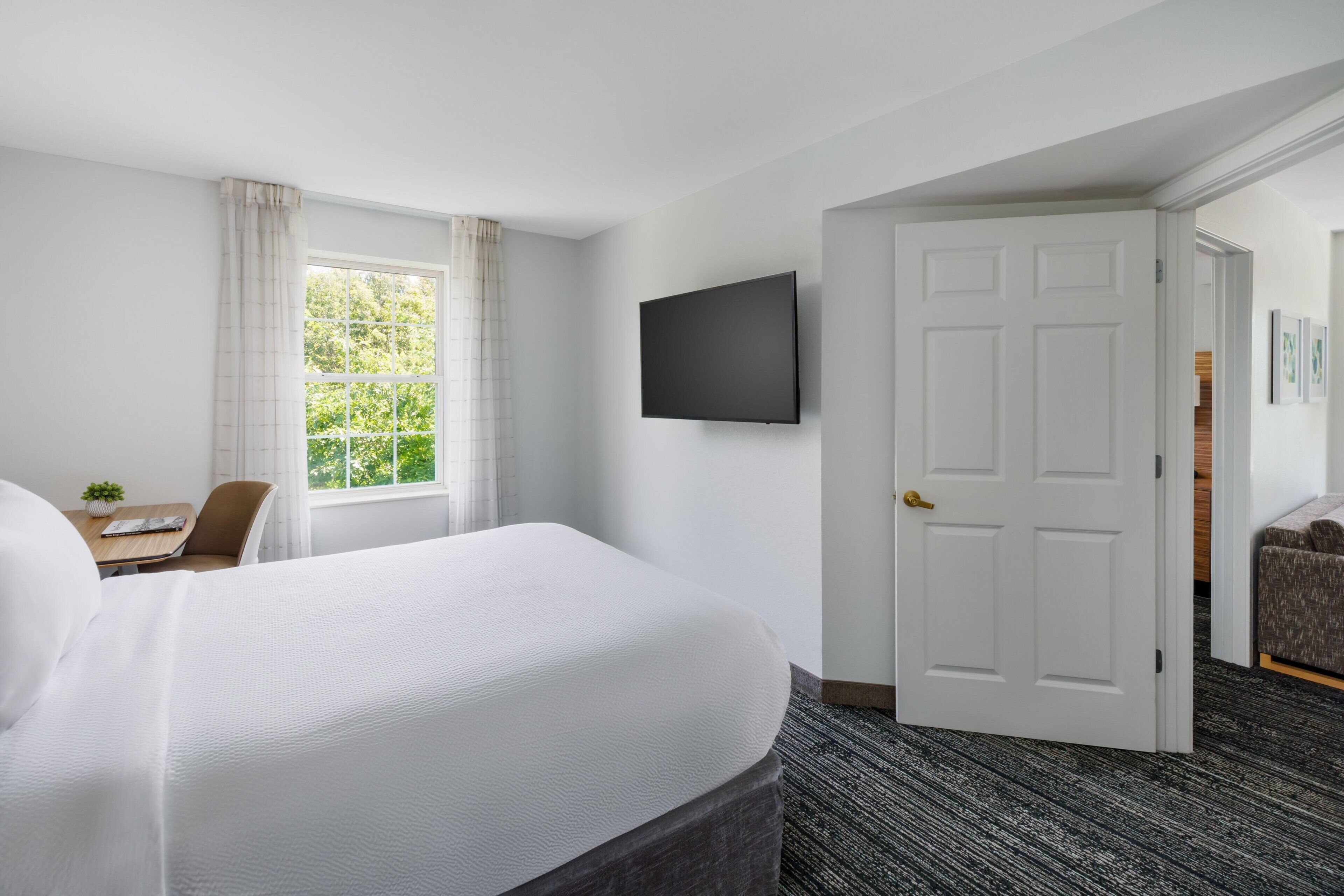 A hotel room with a large bed, a desk with a chair, a television mounted on the wall, and a door leading to another room.