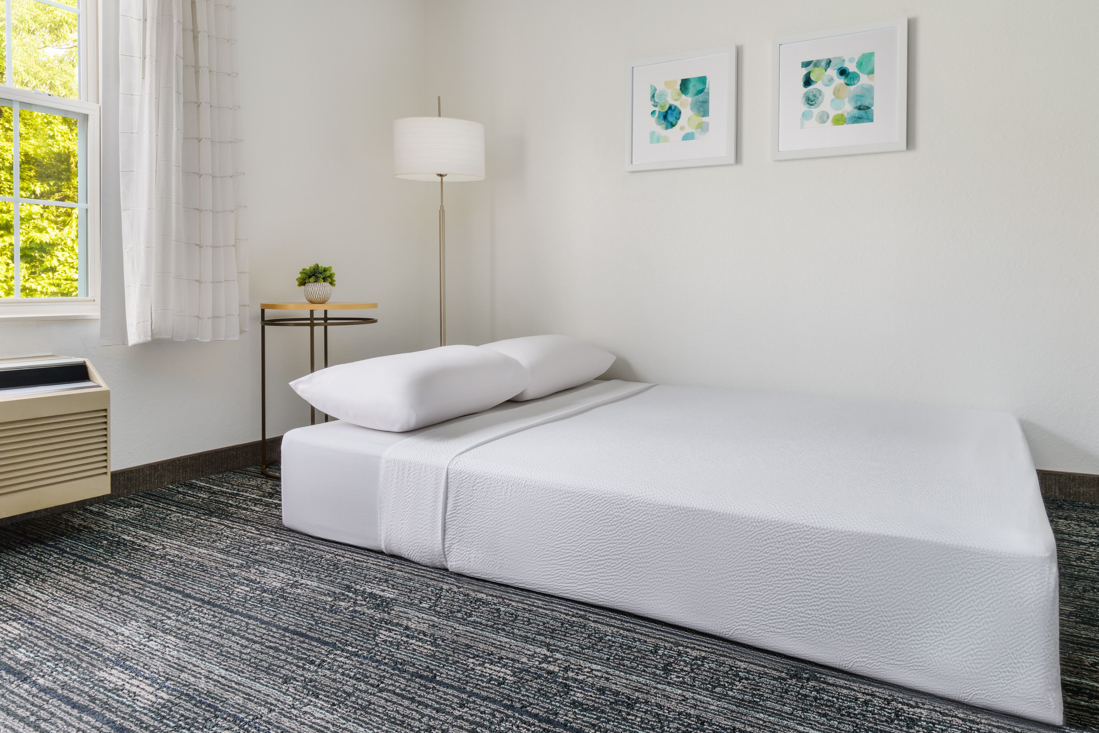 Un dormitorio ordenado con cama, mesita de noche con lámpara y dos cuadros enmarcados en la pared.