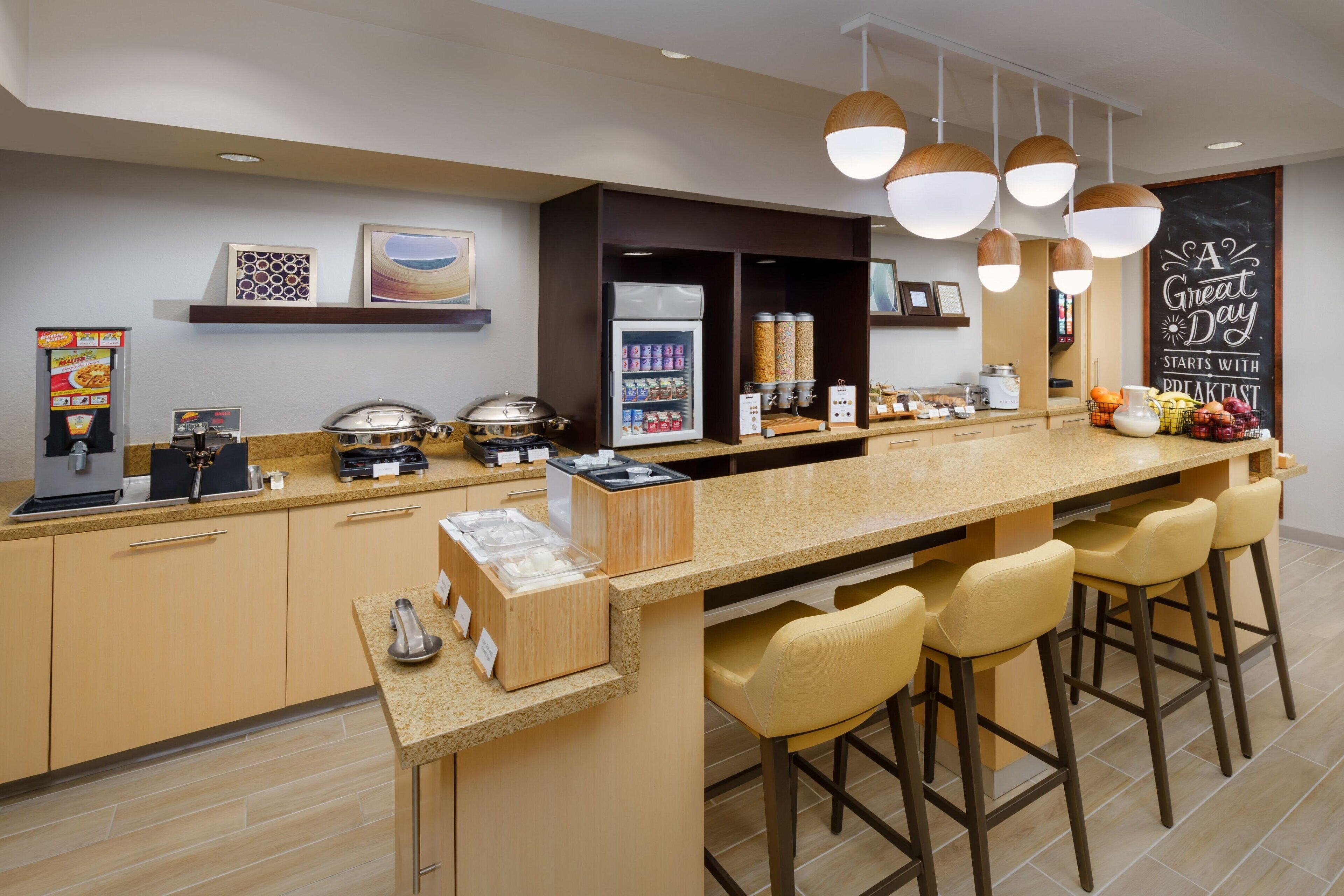 Una cocina moderna con una encimera larga, taburetes de bar, una cafetera y un refrigerador lleno de bebidas.