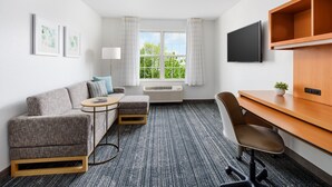 A hotel room with a gray sofa, a small round table, a wooden desk, a chair, a TV, and a window with curtains. - TownePlace Suites by Marriott Manchester (Manchester)