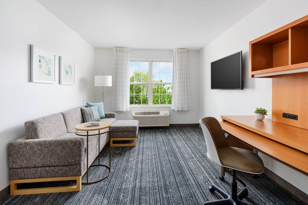 A hotel room with a gray sofa, a small round table, a wooden desk, a chair, a TV, and a window with curtains.