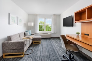 Habitación de hotel con un sofá gris, una pequeña mesa redonda, un escritorio de madera, una silla, un televisor y una ventana con cortinas.