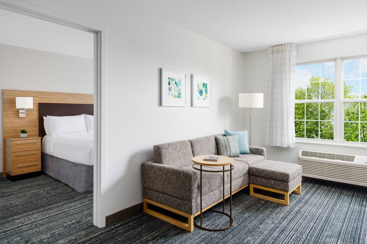 A modern hotel room with a grey sofa, a small round table, a bed with a wooden headboard, and a large window with a view of trees.