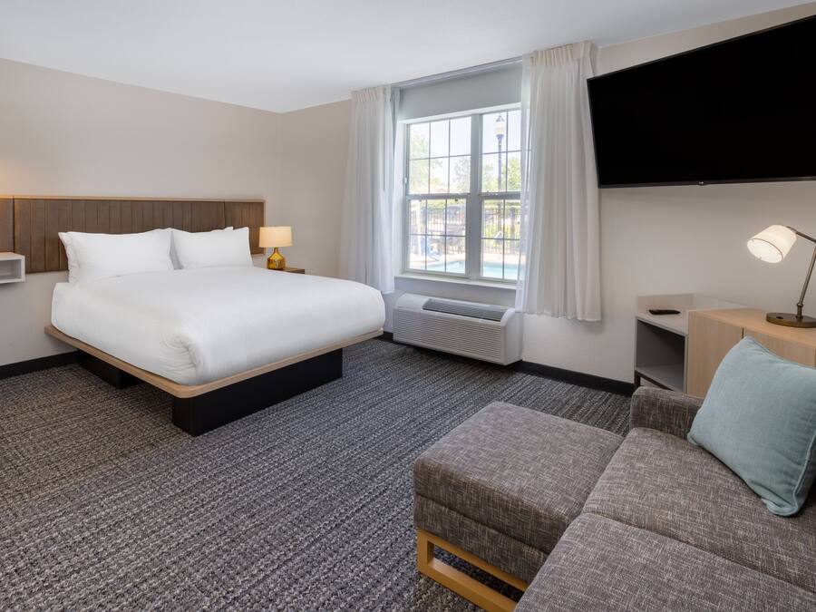 Una habitación de hotel con una cama grande, un sofá, un televisor, una ventana con cortinas y un televisor montado en la pared.