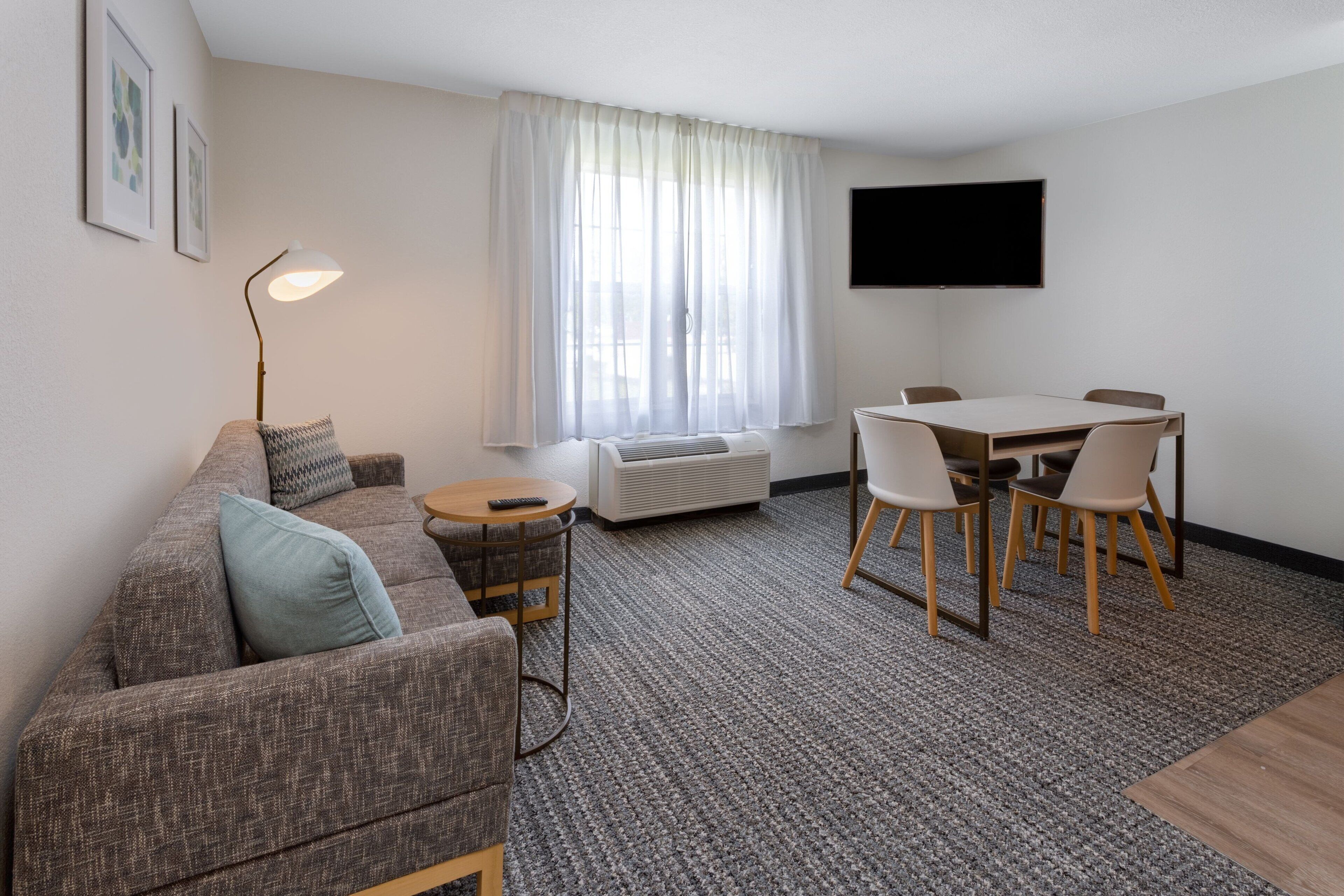 Una sala de estar moderna con un sofá gris, una mesa de centro redonda, una mesa de comedor con cuatro sillas, un televisor de pantalla plana montado en la pared y un amplio ventanal con cortinas translúcidas.