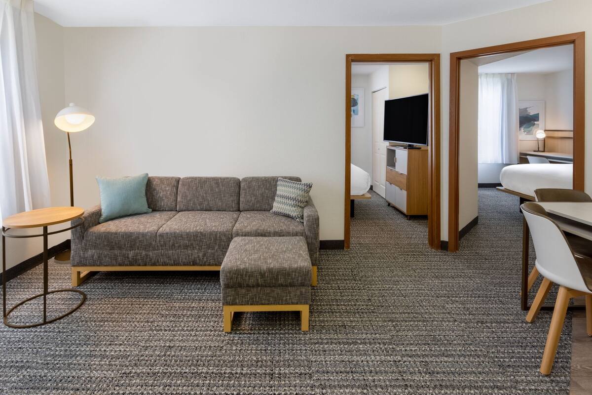 A modern hotel room with a grey sofa, a small table, a lamp, and a TV in the corner.