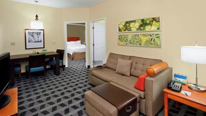 Una habitación de hotel con sofá, reposapiés, escritorio, televisión y obras de arte en las paredes.