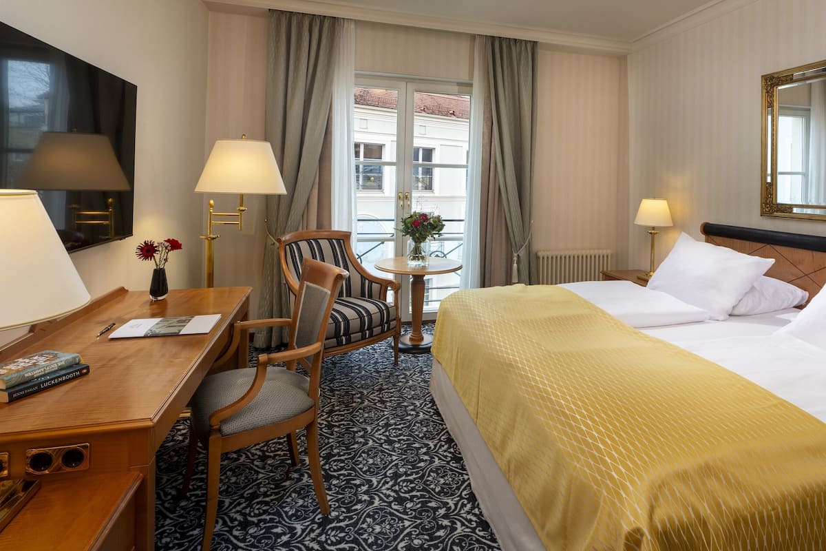 Habitación de hotel con cama, escritorio con lámpara, silla, una mesita con un florero y un espejo grande.