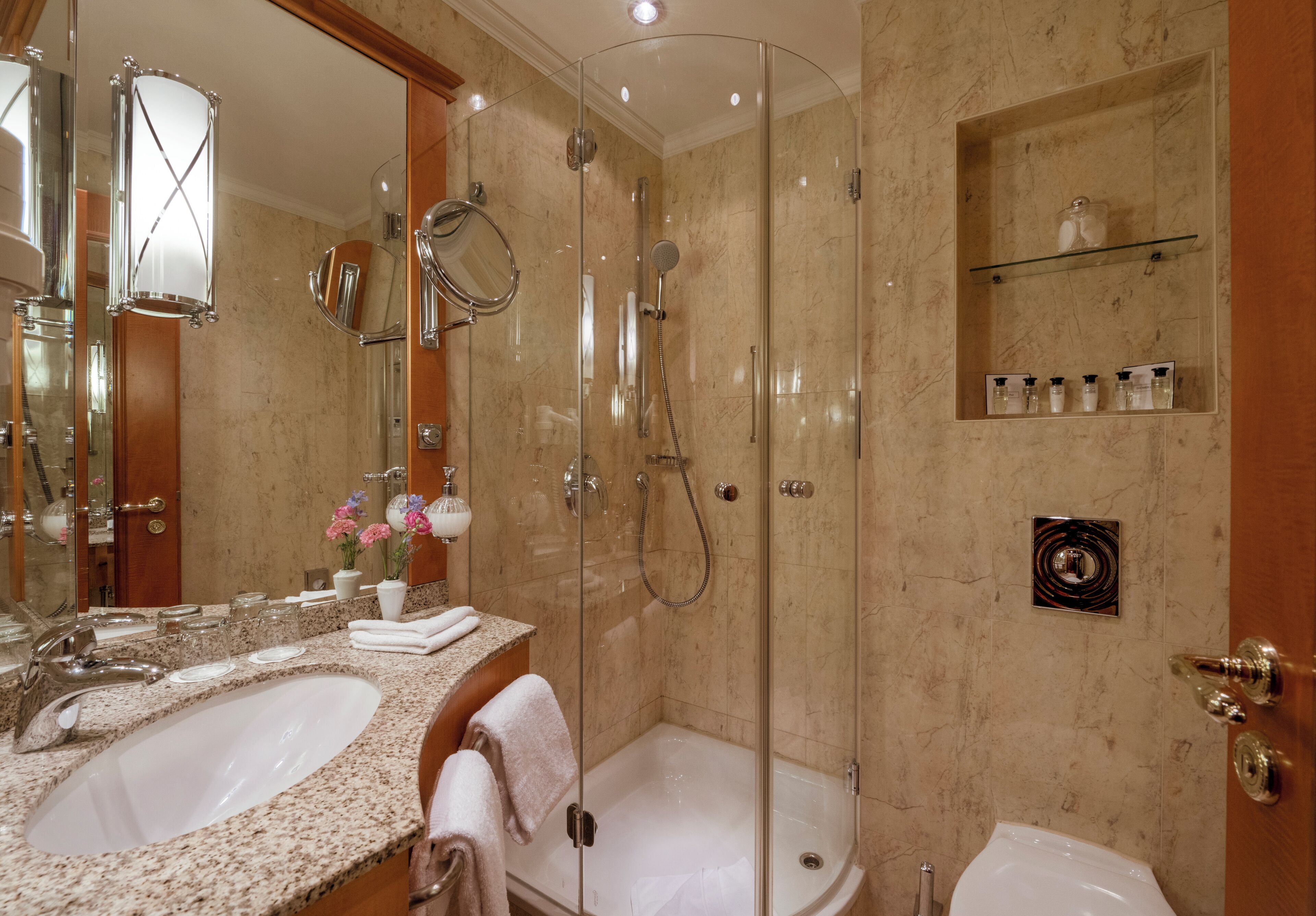 A bathroom with two sinks, a glass-enclosed shower, a toilet, and a shelf with toiletries.