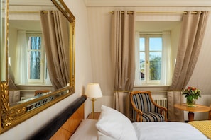 A hotel room with a bed, a chair, a table with a vase, a lamp, and curtains.