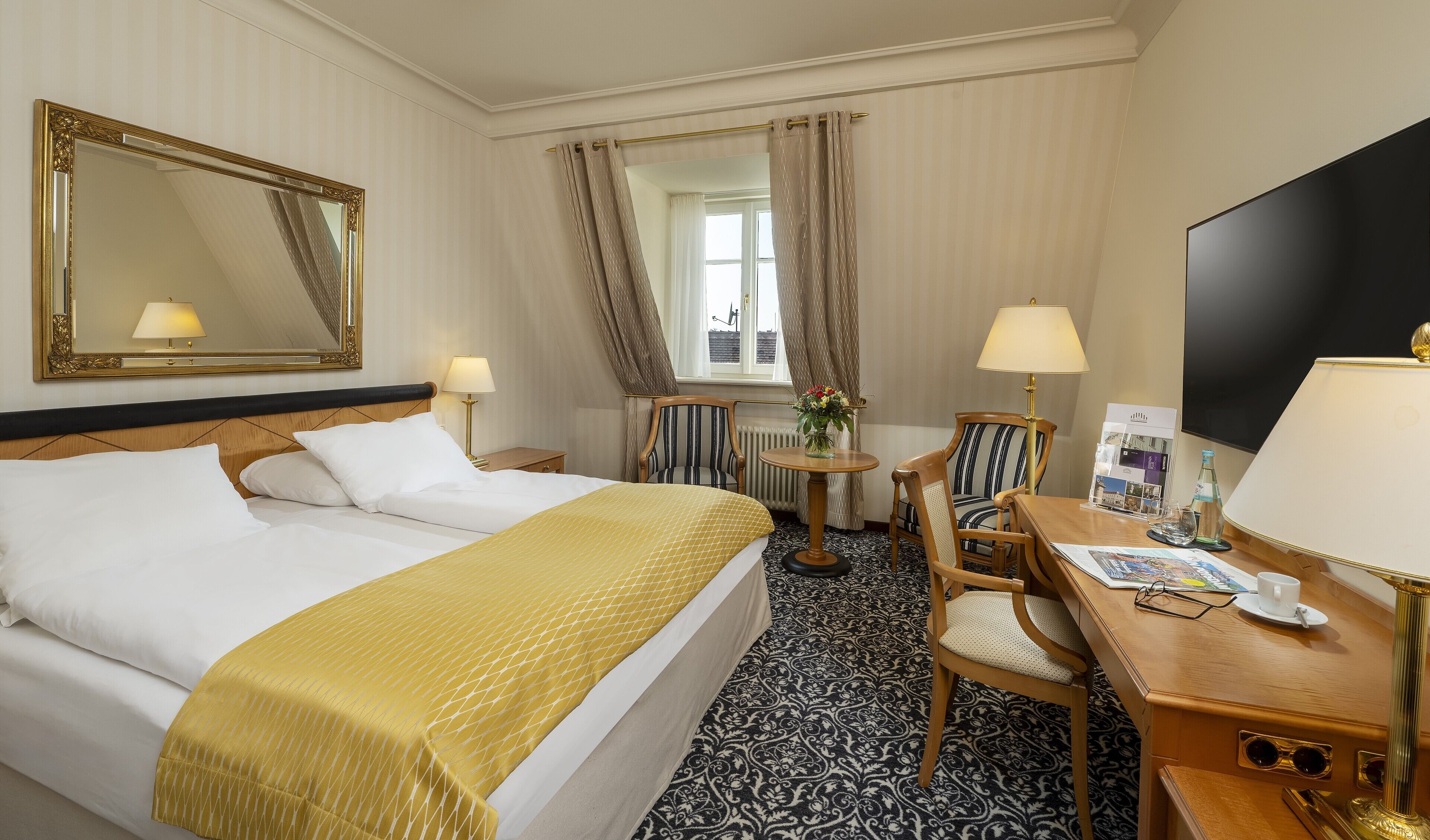 A hotel room with a large bed, a desk with a chair, a TV, and a window with curtains.